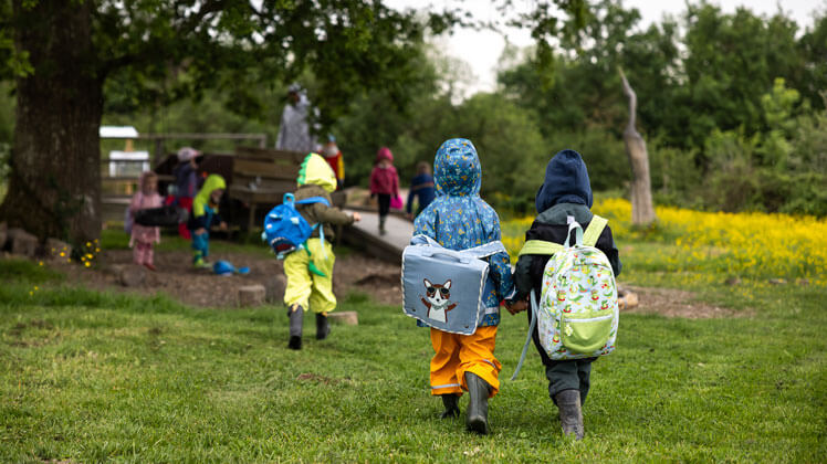 Un groupe de jeunes enfants marche dans un espace naturel herbeux, entouré d’arbres et de fleurs jaunes. Ils portent des vêtements de pluie colorés — vestes, pantalons imperméables et bottes — ainsi que des petits sacs à dos.