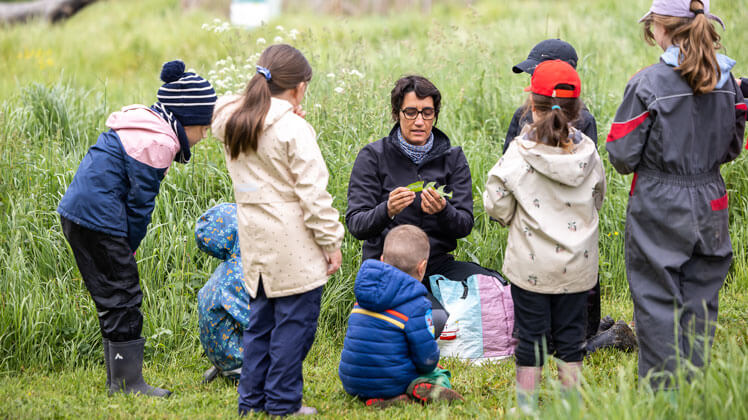 Un groupe d’enfants est rassemblé autour d’un enseignant assis dans l’herbe, dans un espace naturel verdoyant.