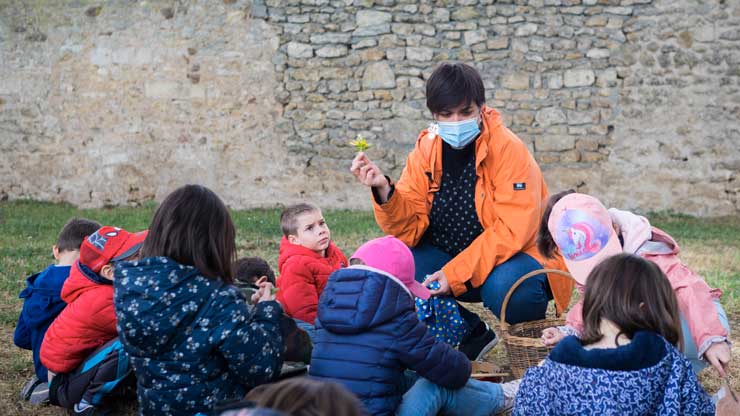 Assis dans l’herbe, autour de leur enseignante, des enfants écoutent attentivement.